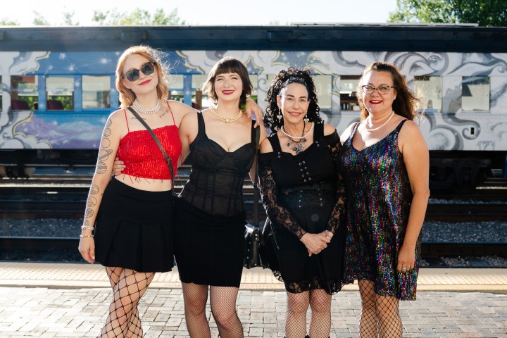 Four women in stylish outfits standing on a platform with a graffiti-covered train behind them.