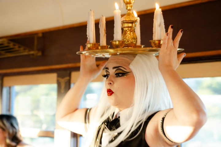 Person with white hair balancing a lit candelabra on their head inside a room.