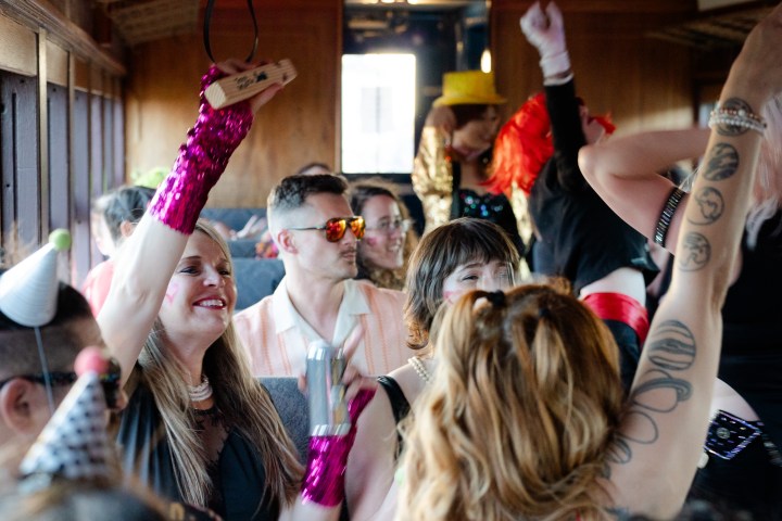People in colorful costumes and hats celebrating on a moving train, raising arms and smiling energetically.