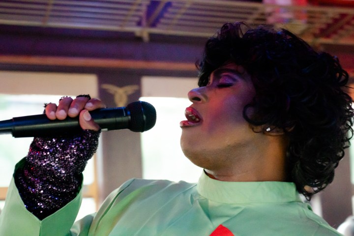Person with curly hair singing into a microphone, wearing sequined gloves and a light green outfit.