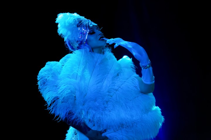 Performer in feathers and headdress posing under blue light on dark stage.