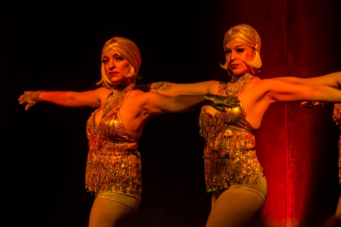 Two performers in glittery costumes and short blonde wigs on stage with dramatic lighting.