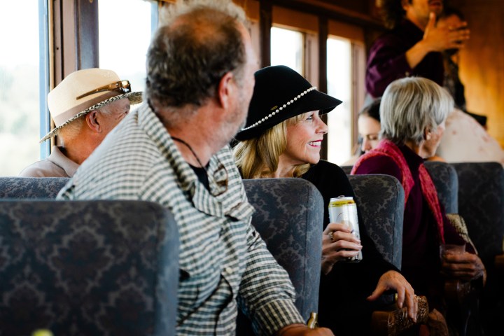 People sitting on a train, one holding a can, wearing hats and smiling.