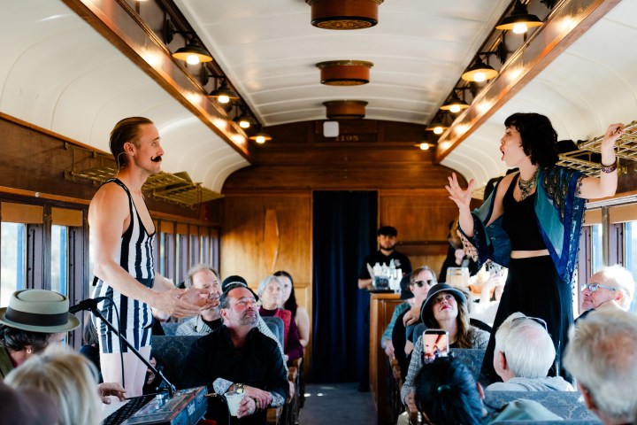 Two performers entertain passengers inside a vintage train carriage.