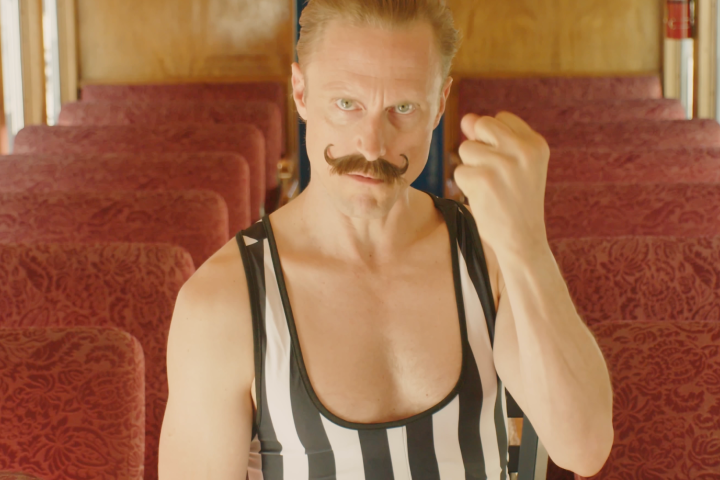 Man with a mustache in a striped outfit, raising fist in a vintage train car.