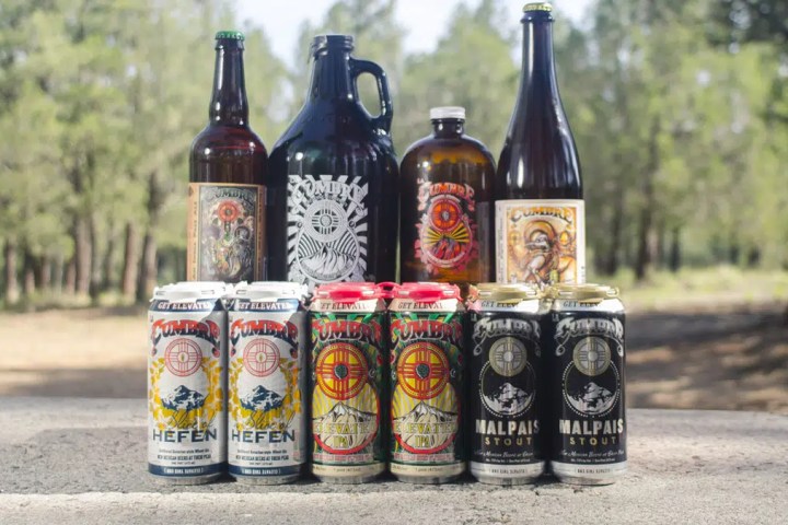 Assorted beer bottles, cans, and a growler on a table outdoors with trees in the background.