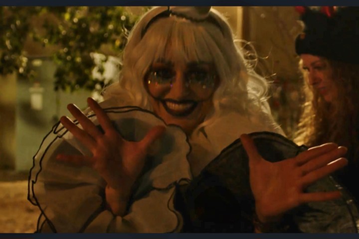 Person in spooky clown costume with white wig and large ruffled collar, hands raised, another person in background.
