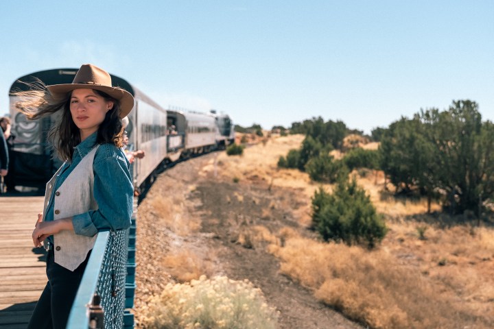 Woman in hat stands on bridge next to moving train in rural landscape.