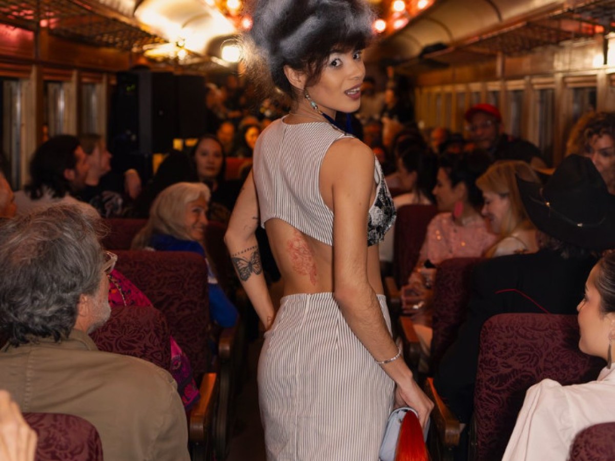 Person in striped dress with big hair, walking down a crowded train aisle, looking back at the camera.