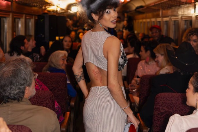 Person in striped dress with big hair, walking down a crowded train aisle, looking back at the camera.