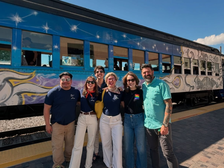 Group of six people smiling in front of a colorfully painted train under a clear blue sky.