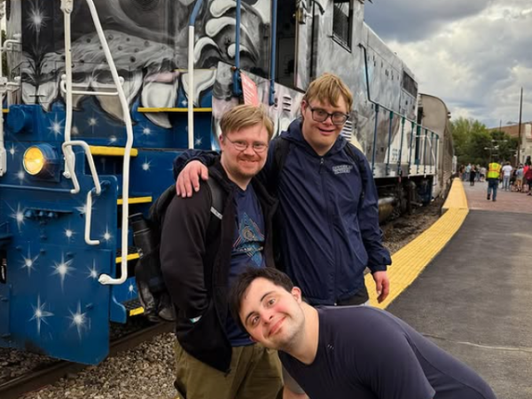 Three people smiling in front of a parked train with artistic decorations.