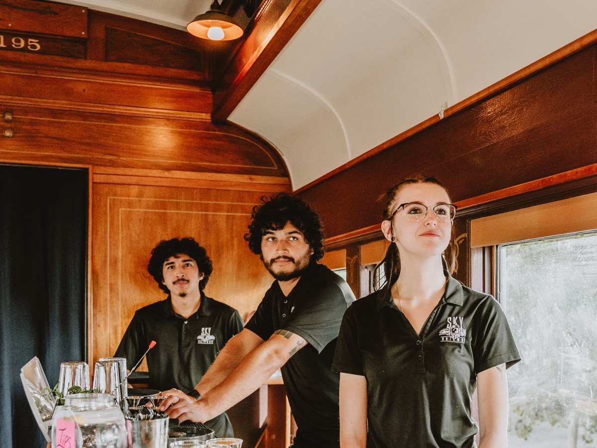 Three bartenders in black uniforms stand behind a bar inside a wooden train car.