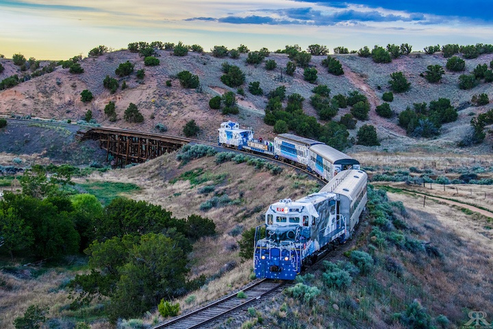 Blue train on curved track in hilly landscape with sparse vegetation and cloudy sky.
