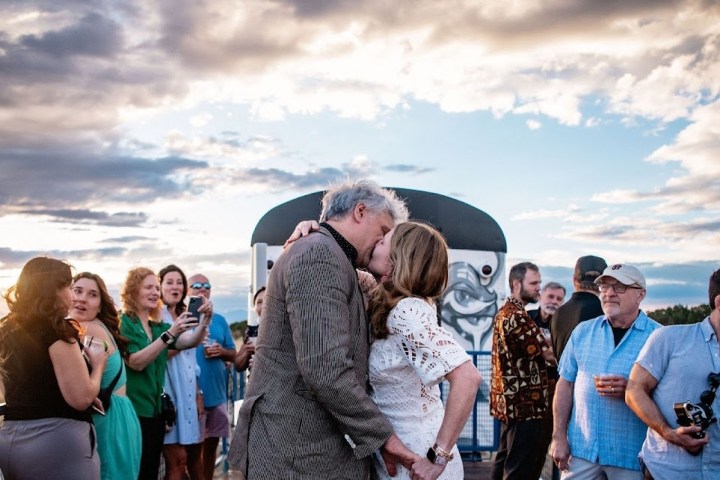 Couple kissing at an outdoor gathering with people around, dramatic sky background.
