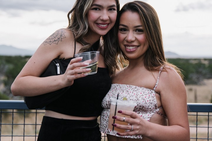 Two smiling women holding drinks, standing outdoors by a fence.