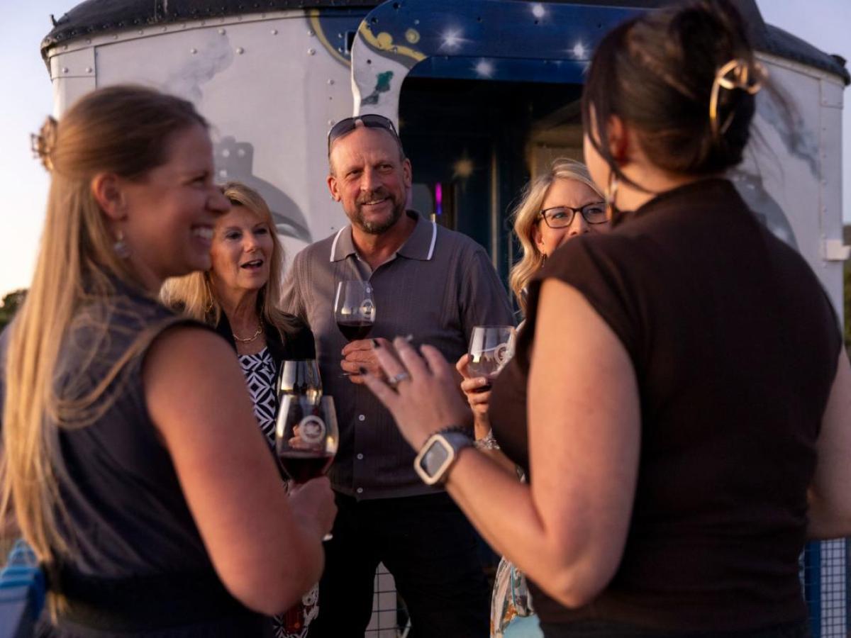 Group of people smiling and drinking wine outdoors near a trailer.