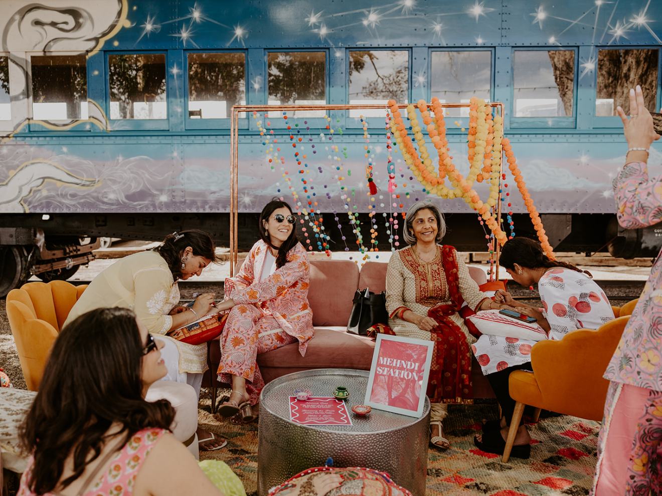 Women sitting by mehndi station with colorful decorations and train backdrop.