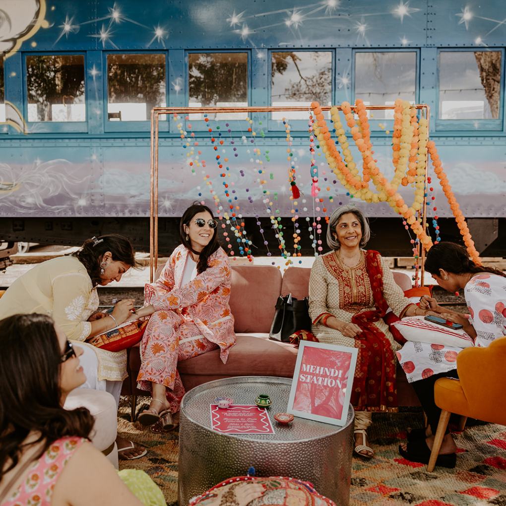 Women sitting by mehndi station with colorful decorations and train backdrop.