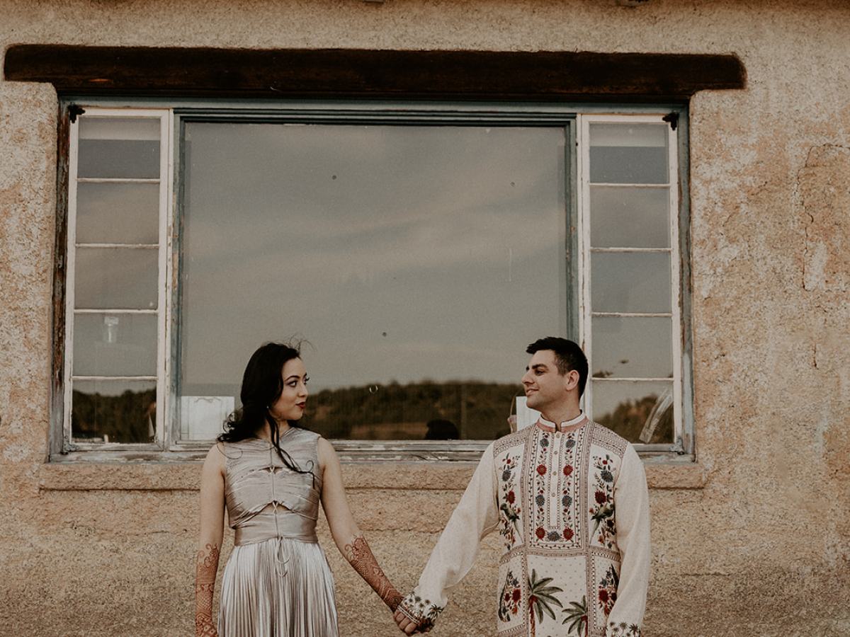 A couple in traditional attire holding hands in front of an old building with 'LAMY' sign.