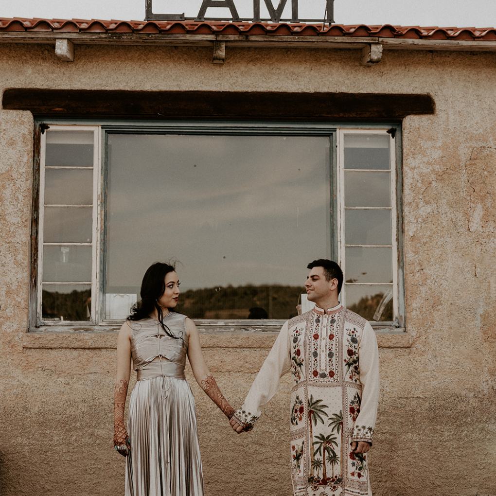 A couple in traditional attire holding hands in front of an old building with 'LAMY' sign.
