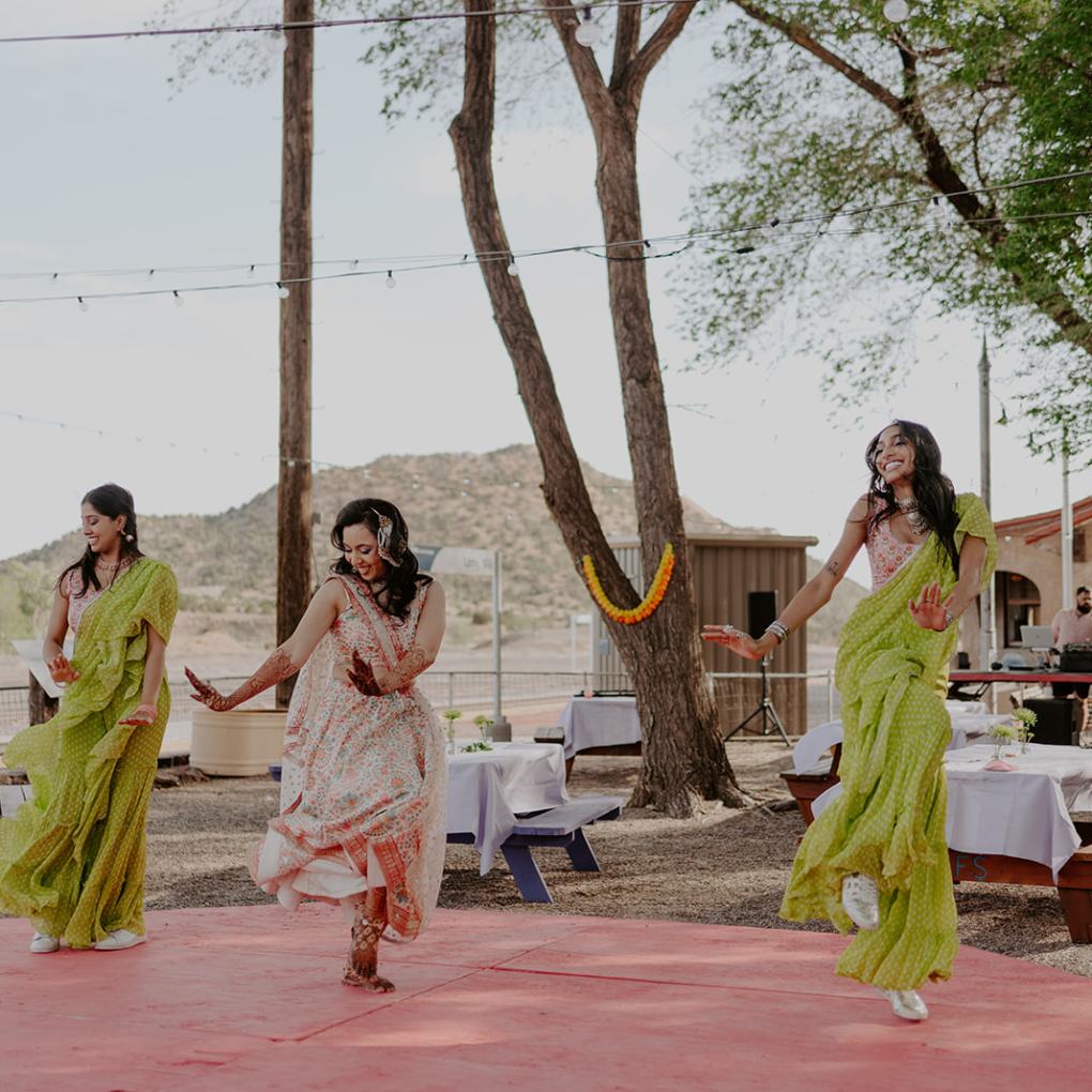 Three women dance outdoors on a circular stage with picnic tables and trees around.