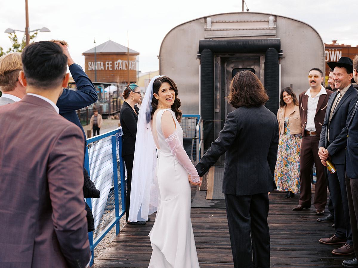 Bride and groom holding hands, surrounded by guests on a wooden platform.