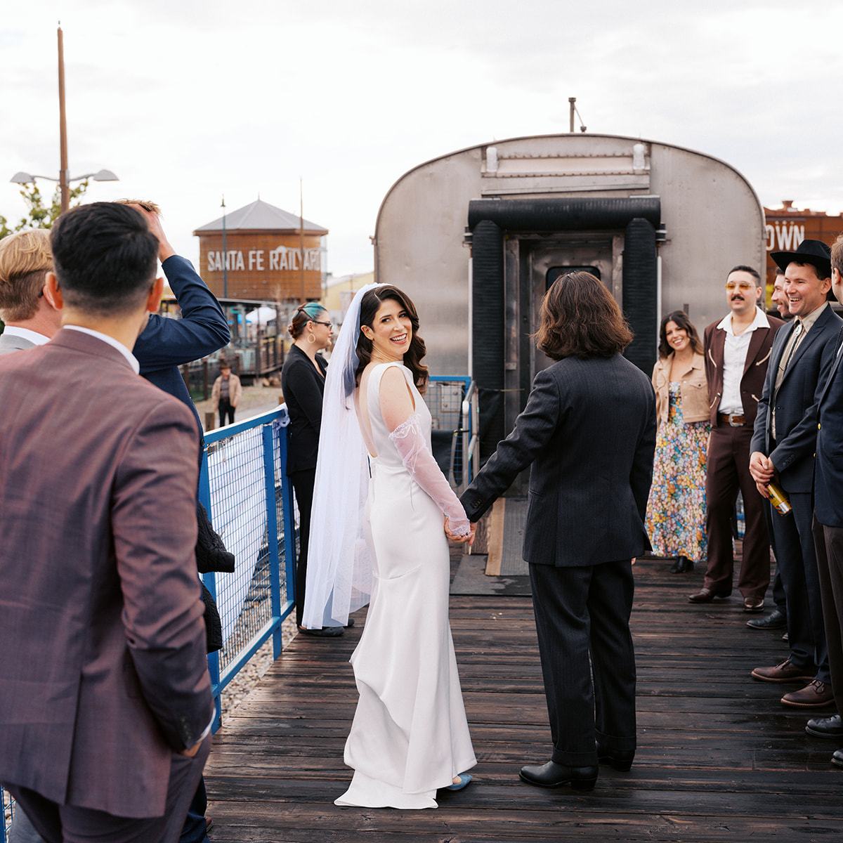 Bride and groom holding hands, surrounded by guests on a wooden platform.