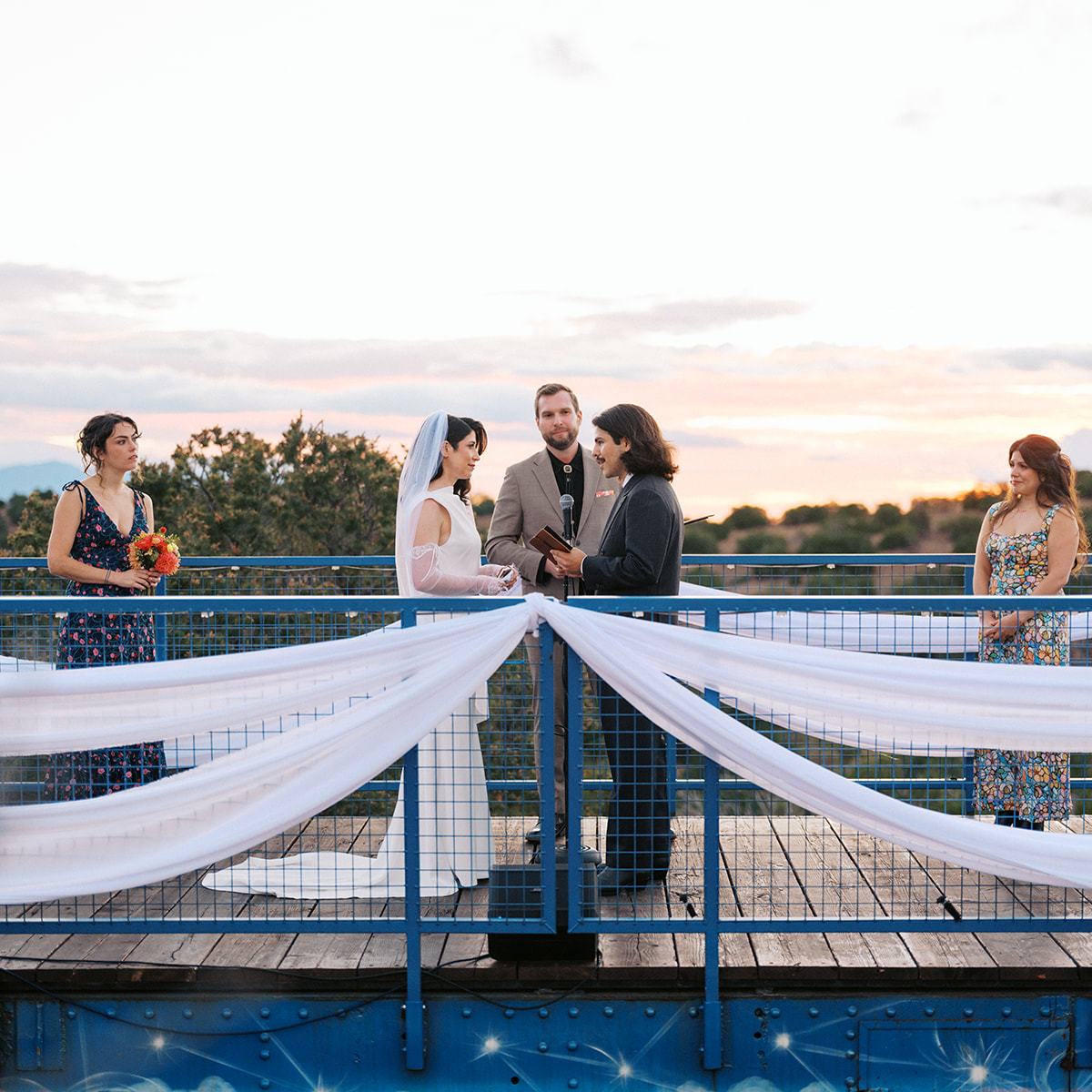 Outdoor wedding ceremony on a bridge with four people, two exchanging vows.