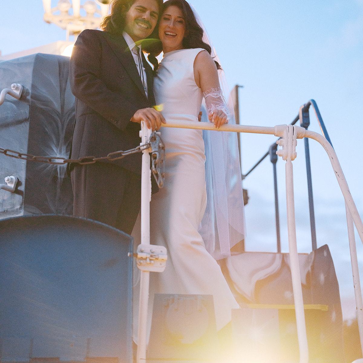 Couple in wedding attire pose on a train with a lit headlight and blue sky backdrop.