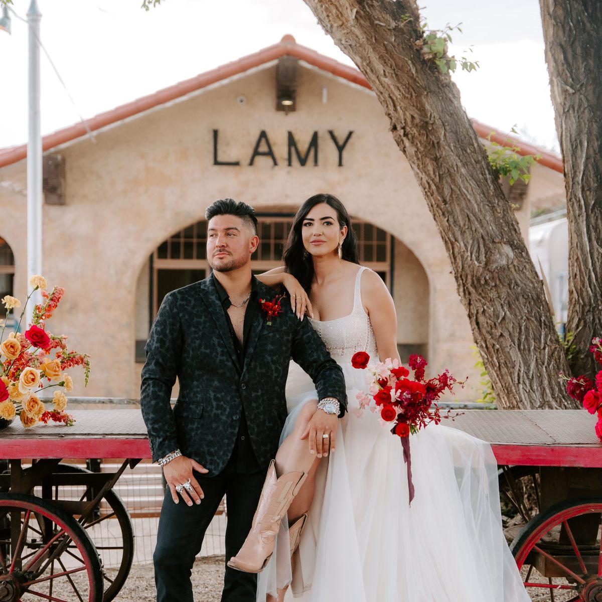 Couple poses with flowers in front of Lamy building; woman in dress and boots, man in dark suit.