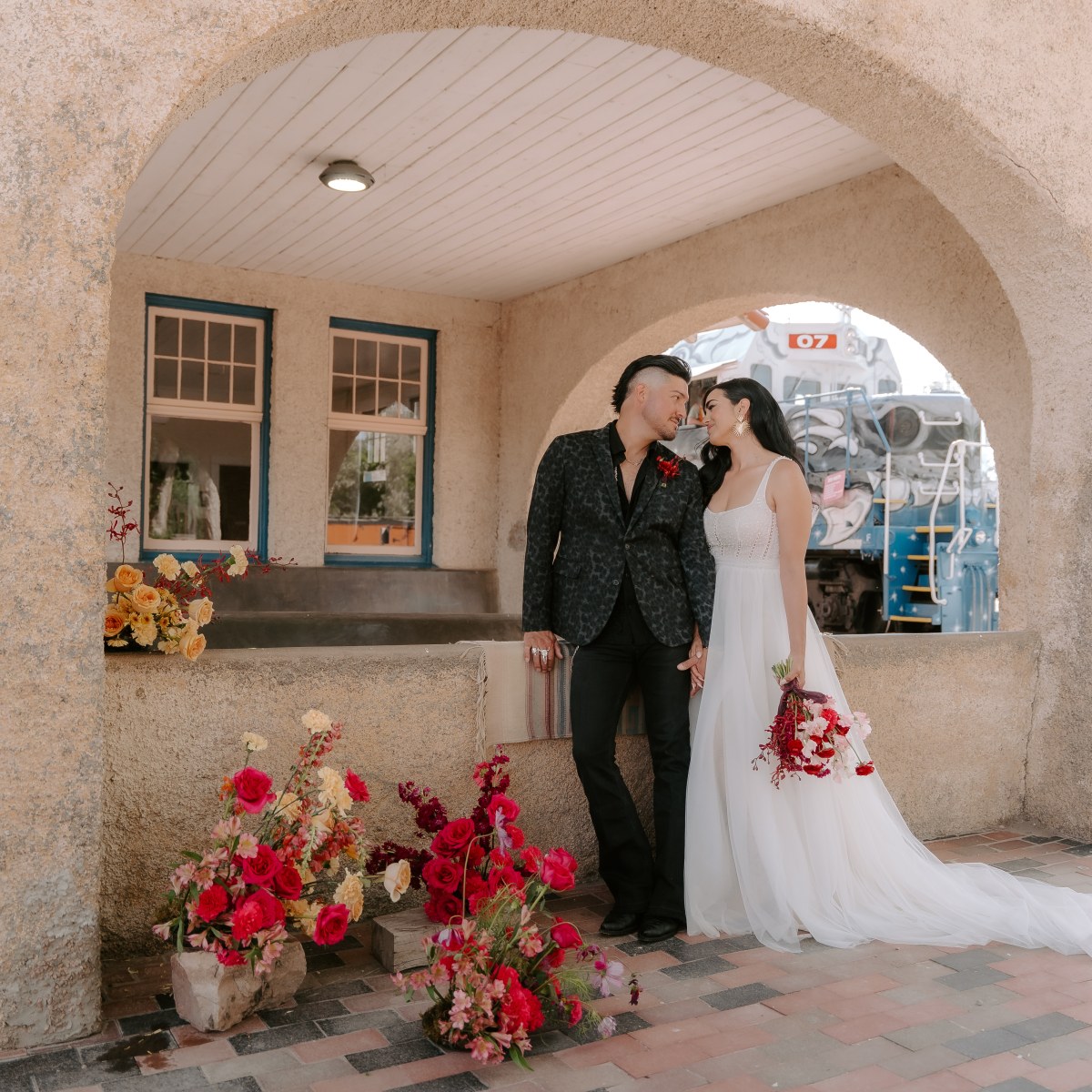 Couple in wedding attire kiss under arched structure near train, with red flowers on the ground.